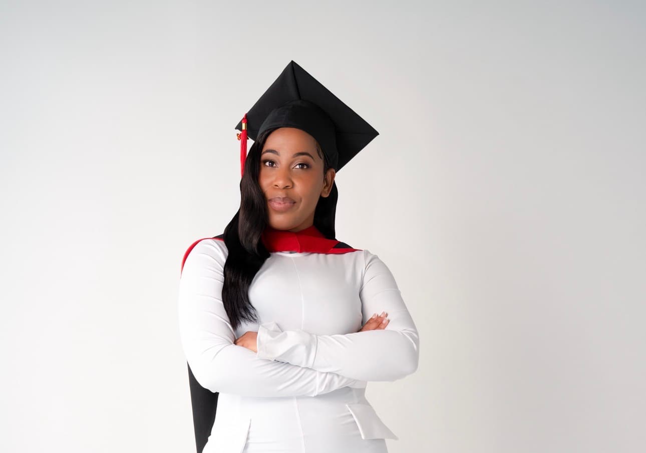 Confident Black woman graduate in a mortarboard and white dress posing with arms crossed.